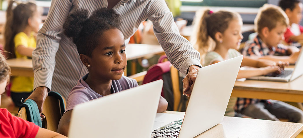 Five kindergartners are working on computers, a teacher helps one of the students