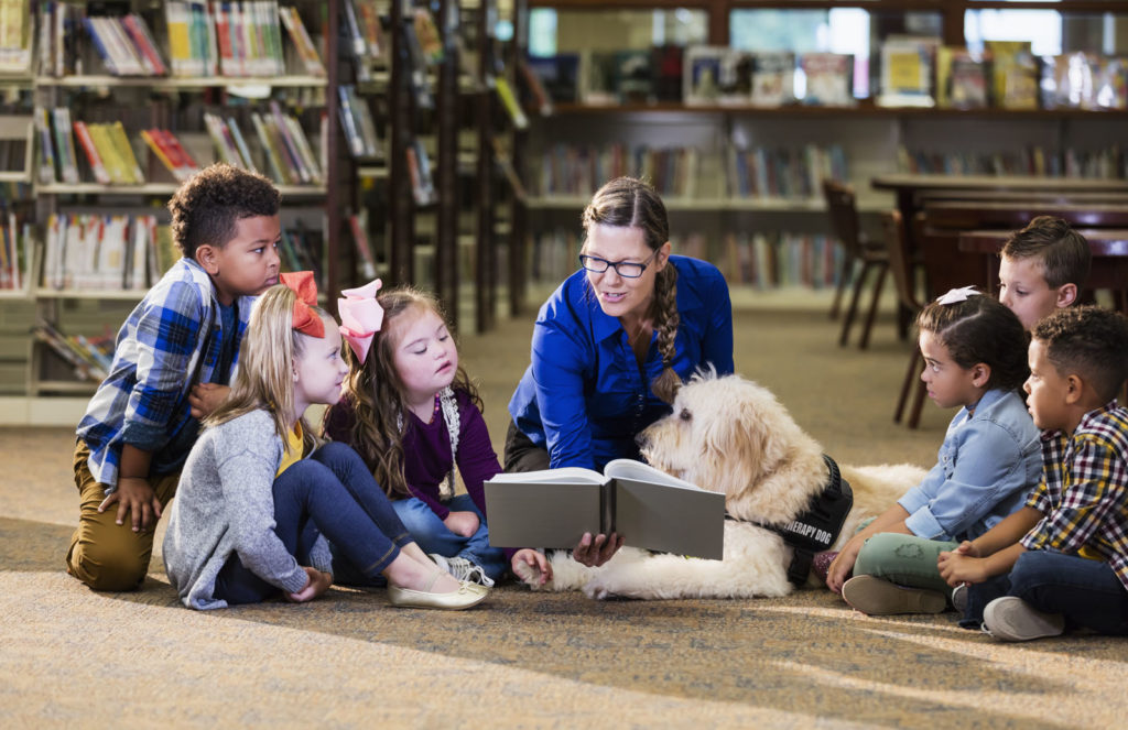 A group of six boys and girls and a woman librarian are sitting on the floor of a library, reading with a therapy dog. 