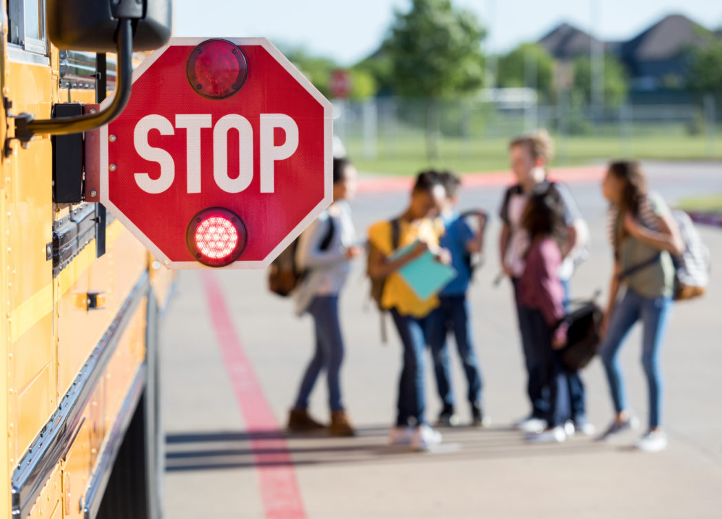 A stop sign is attached to a bus, it is the focus, in the background, middle school students are standing near the bus