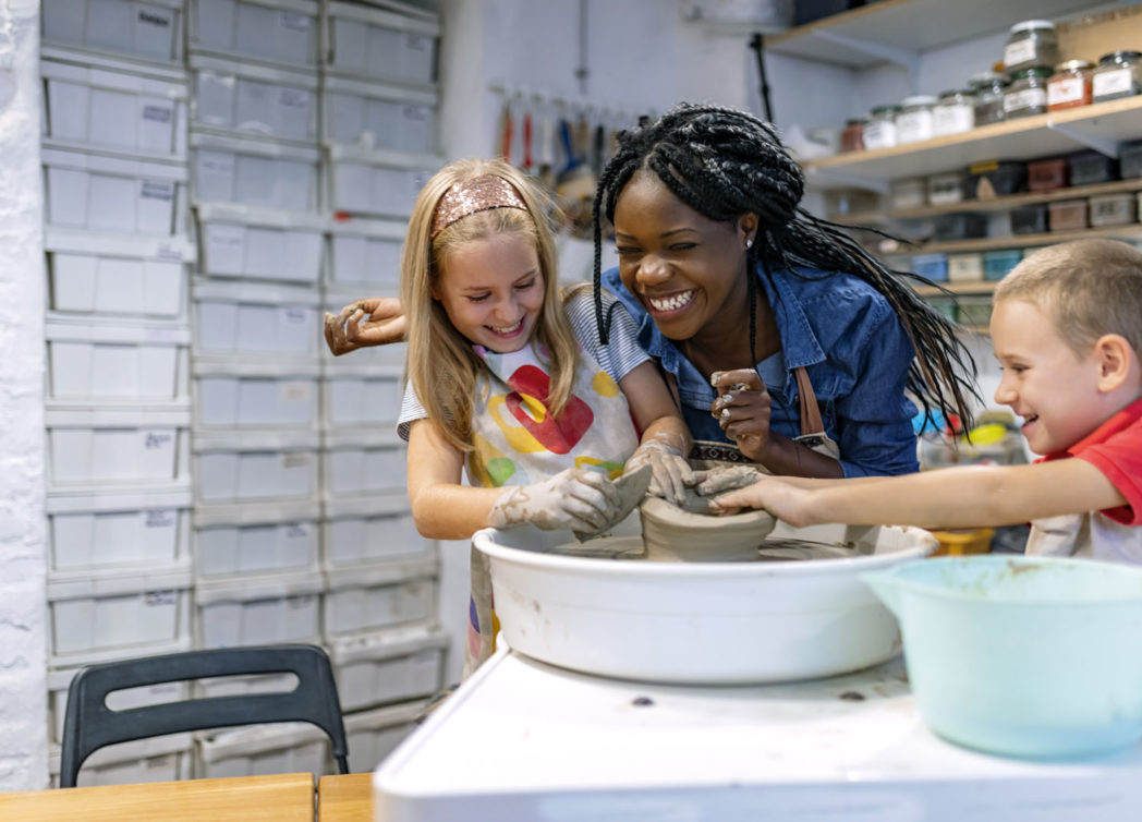 Mother and two children smile inside of a pottery workshop as they sculpt a clay pot